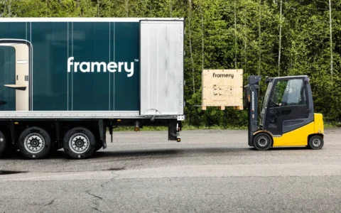 A truck trailer sits on the road beside a yellow forklift and a wooden crate in a forested area.