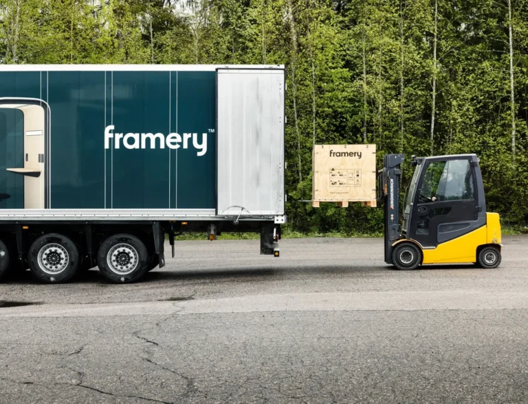 A truck trailer sits on the road beside a yellow forklift and a wooden crate in a forested area.
