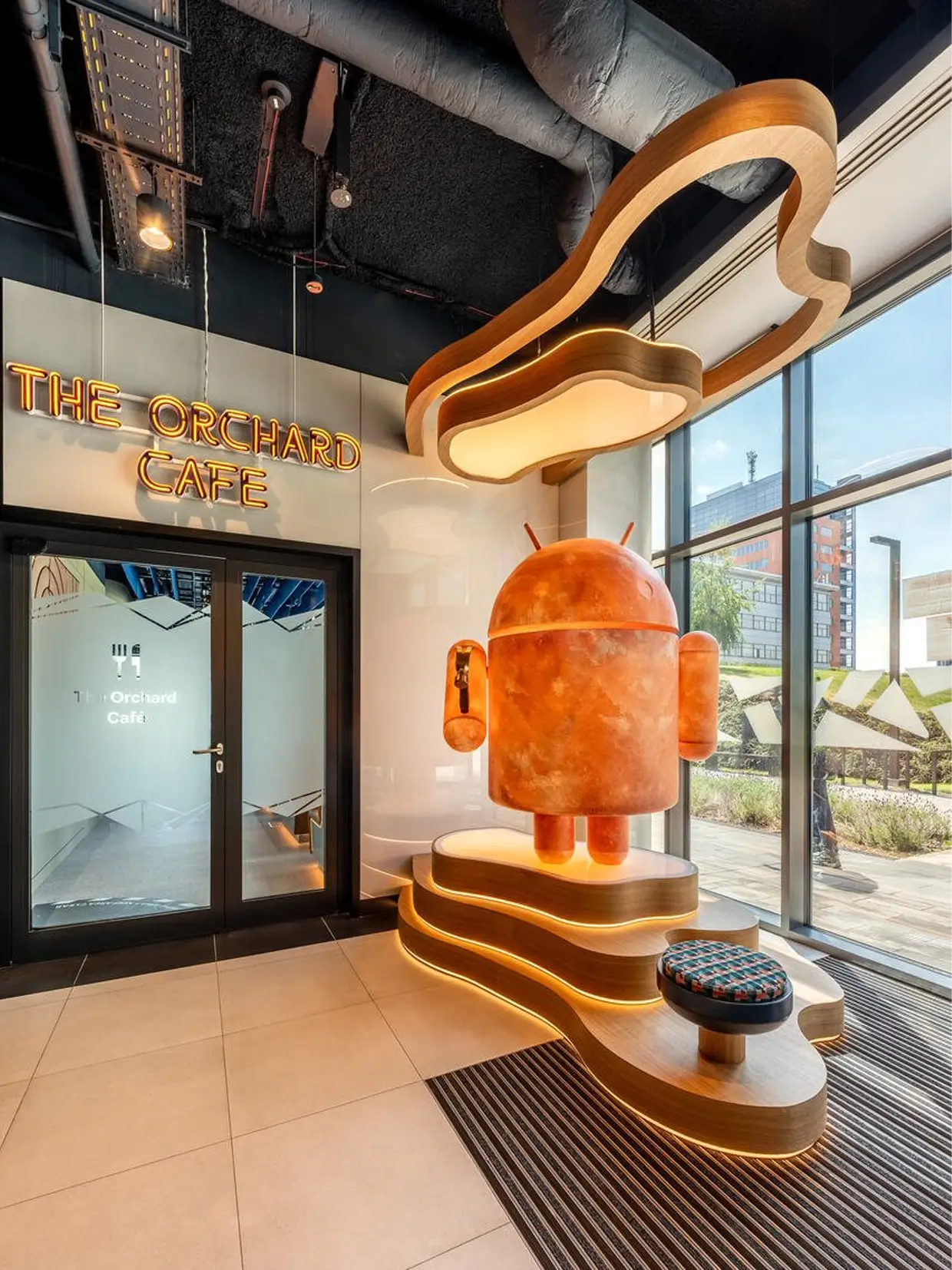 Outside The Orchard Cafe, an orange Android mascot statue stands on a curved, multi-tier wooden pedestal beneath a large round-shaped sculpture.