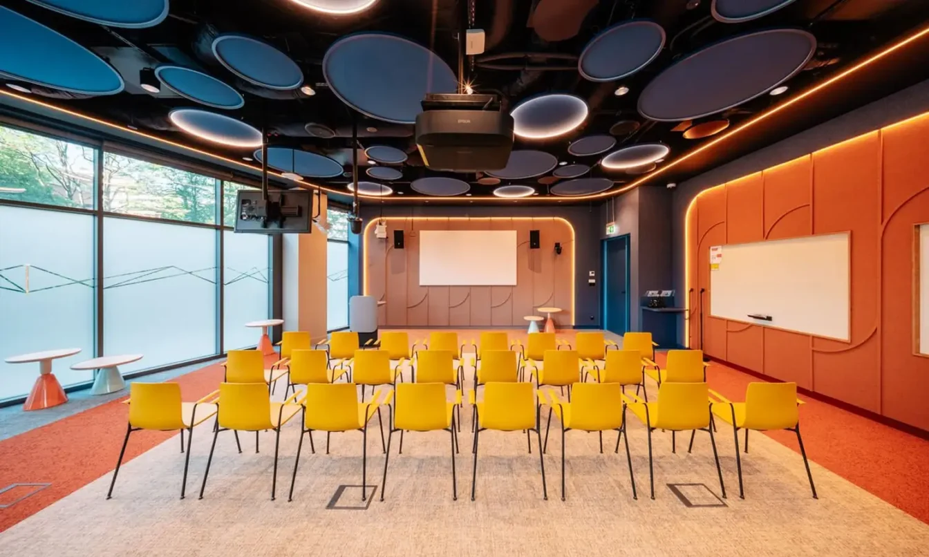 A modern conference room with yellow chairs arranged in rows facing a stage and screen, blue and orange walls, and large windows.