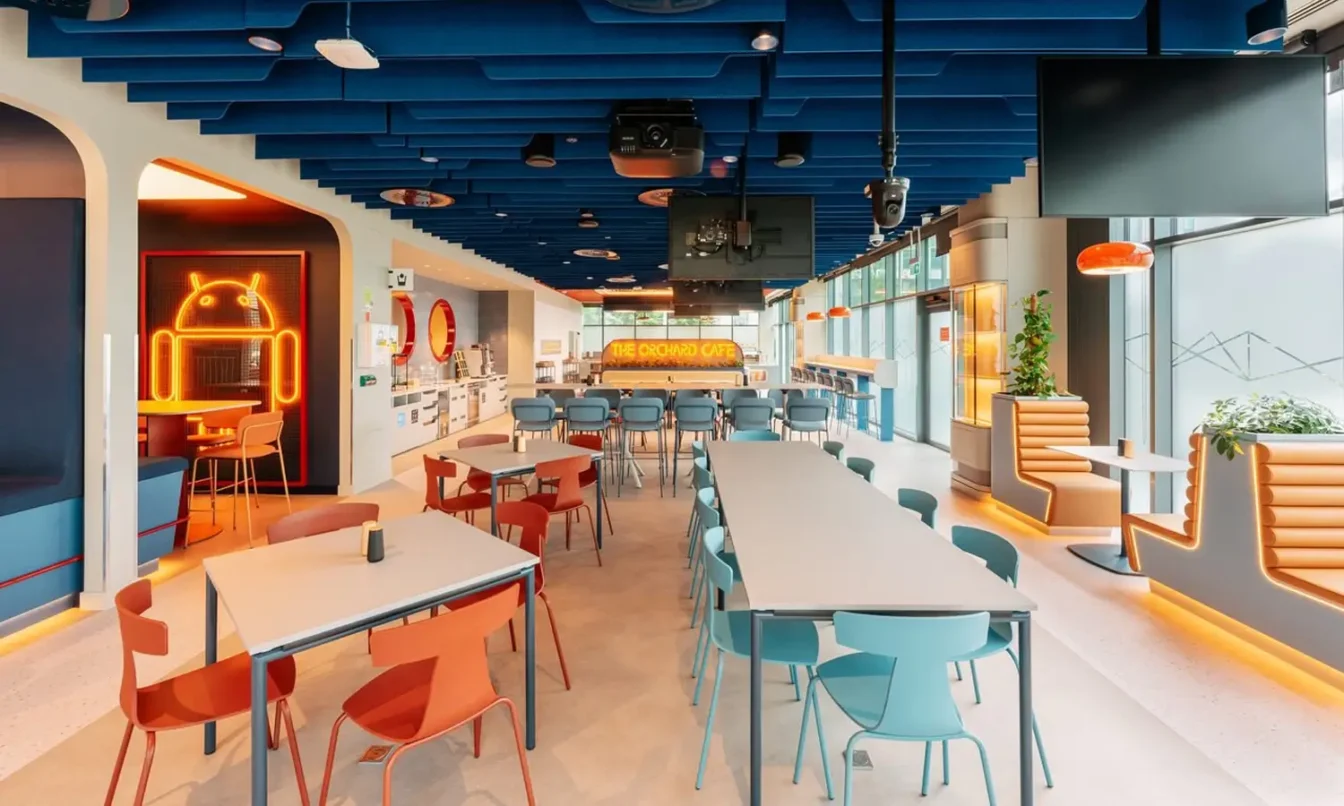 Bright, modern cafeteria with a blue ceiling, long rows of tables, and orange and teal chairs near large windows.