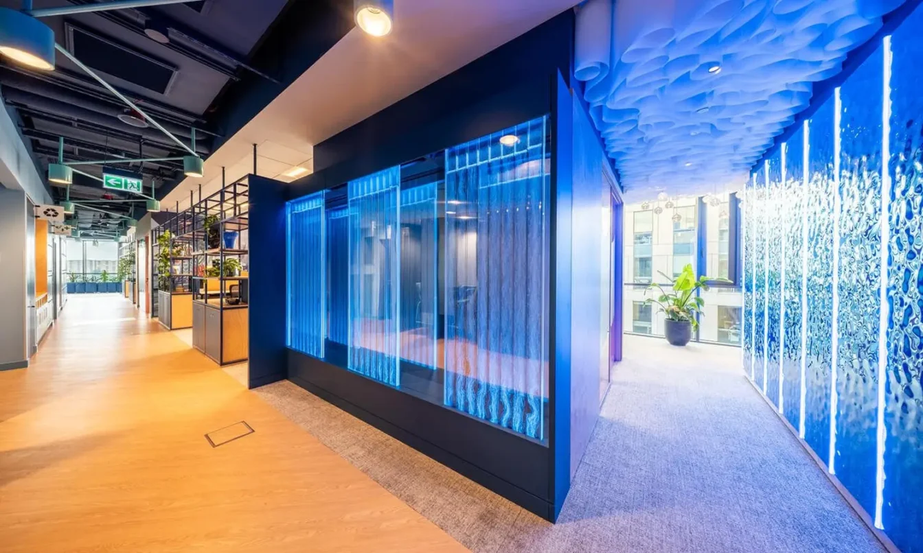 Modern office corridor with blue-toned glass walls on the right, an orange-toned wooden floor on the left, and bright overhead lighting.