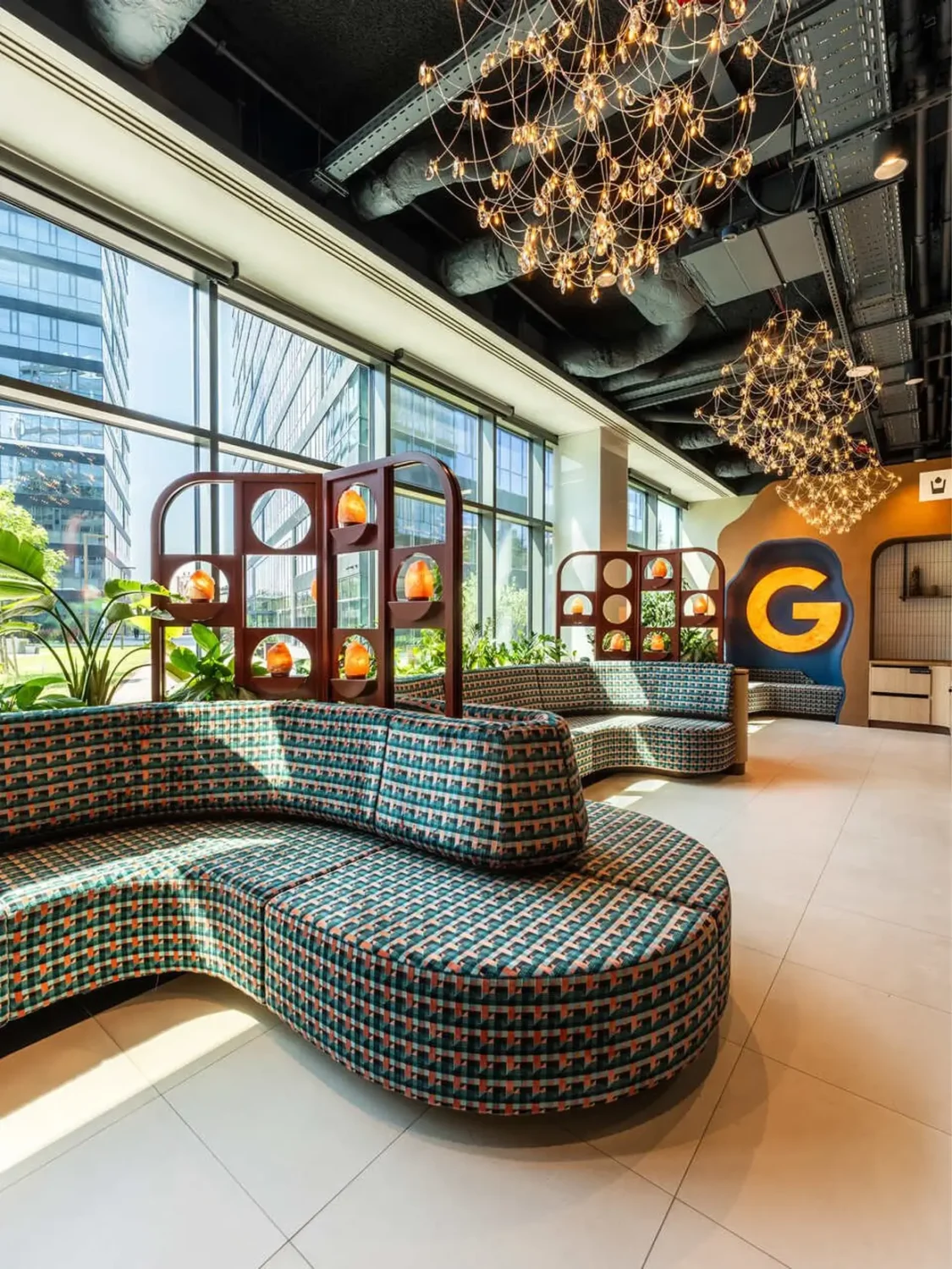 Modern lounge with curved, patterned seating, glass partition shelves, hanging globe chandeliers, and a large G on a yellow wall by expansive windows.